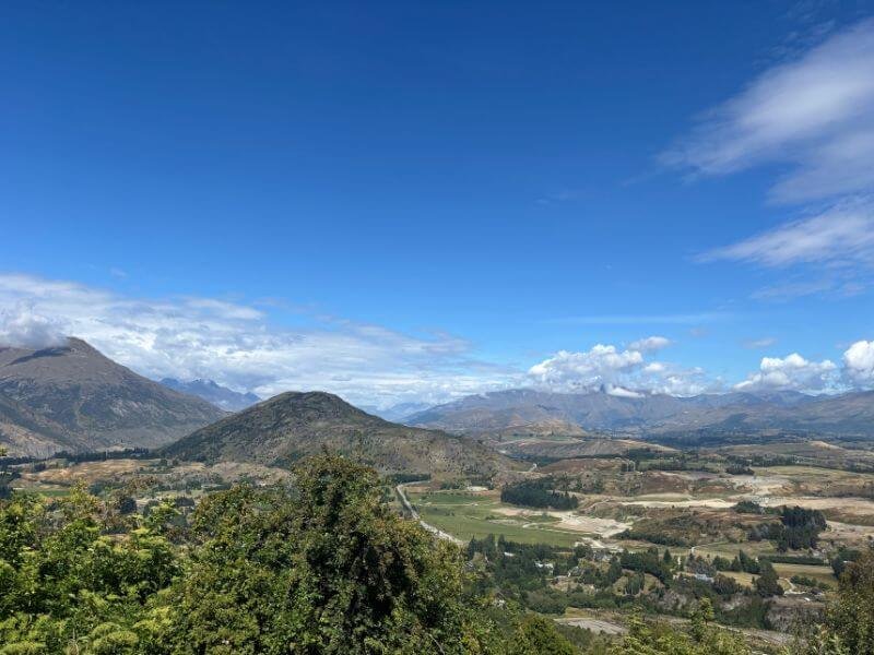 Panoramic mountain views from the Mount Iron Track day hike in Wanaka. Rolling hills, valleys, and distant peaks stretch under a bright blue sky with scattered clouds.